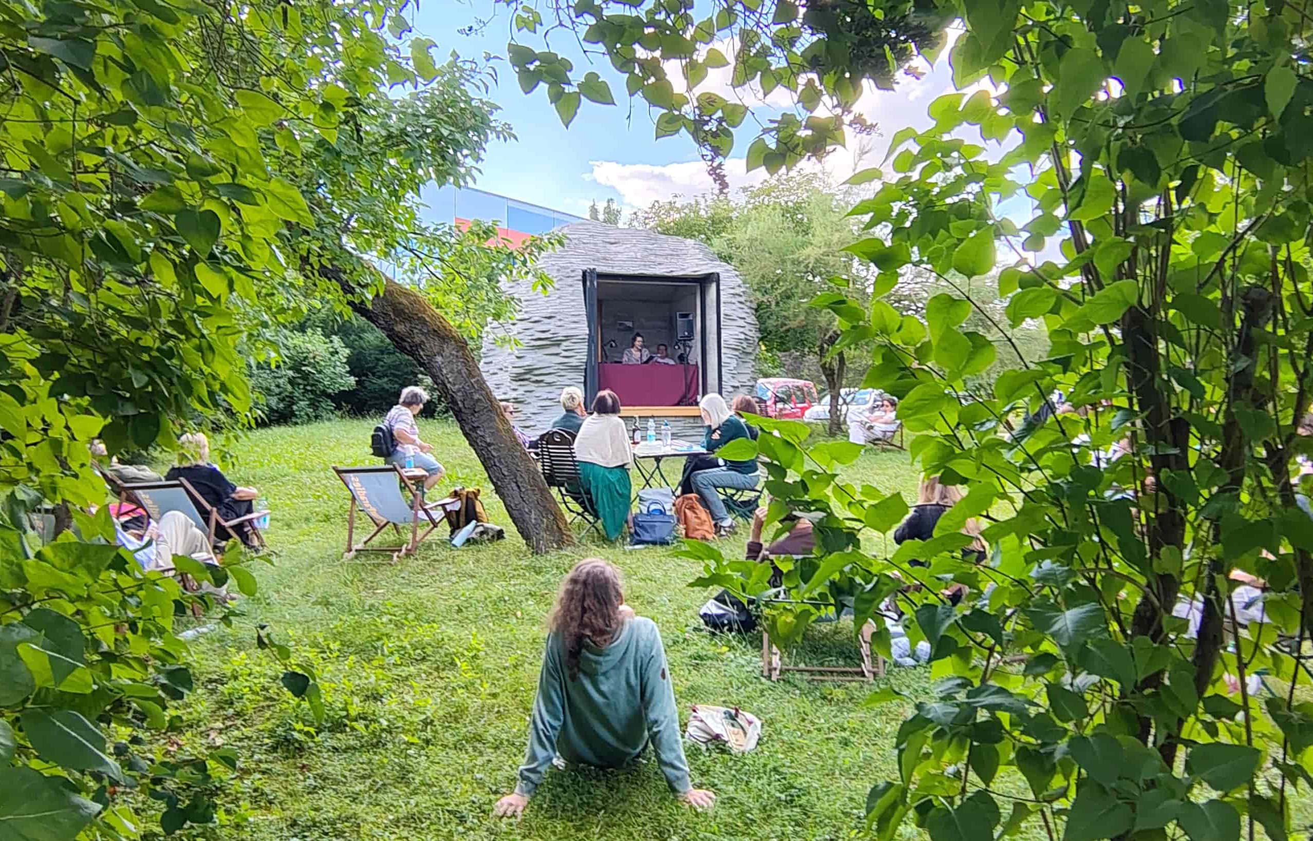 Bühne im Garten davor Menschen in Liegestühlen und auf Picknickdecken