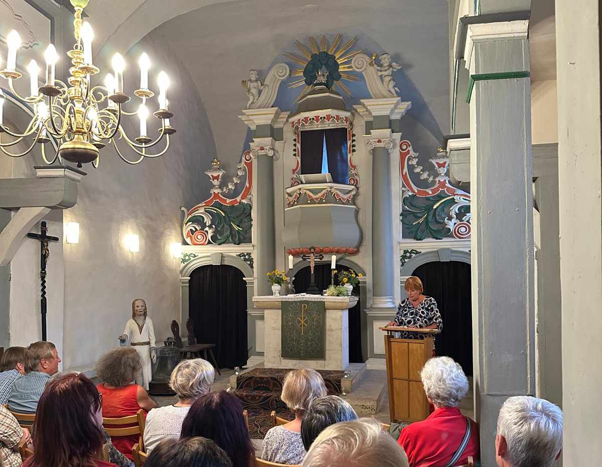 Frau steht am Rednerpult in einer gut gefüllten Kirche mit weiß-grün-rotem Altar